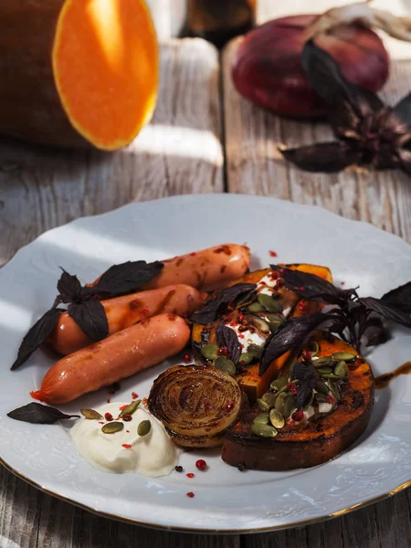 Grilled pieces of pumpkin, onion and sausage on a white ceramic plate. Creamy and balsamic sauce. Decorated with pumpkin seeds and basil. Wooden vintage surface. Cutlery. Close-up. Vertical frame.