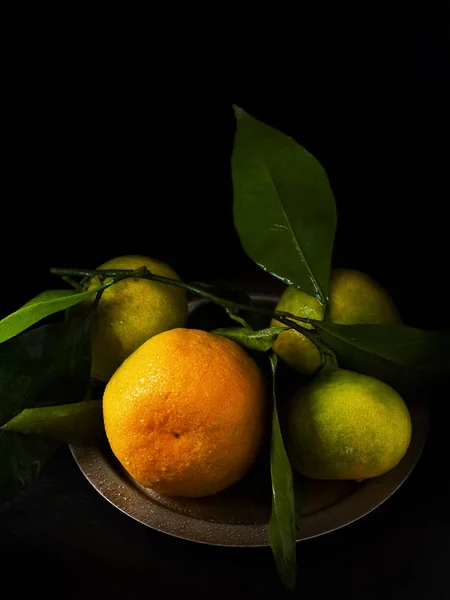 Tangerines with leaves in a rustic metal plate, on a black stone surface. Close-up. Citrus.