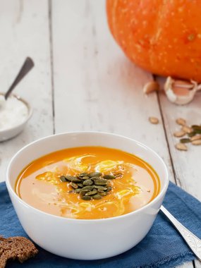 Mavi keten peçeteye kabak çorbası, sos ve kabak çekirdeği konulmuş beyaz kase. Arka planda sos, balkabağı ve sarımsak. Yakın plan. Seçici odaklanma. Cadılar Bayramı