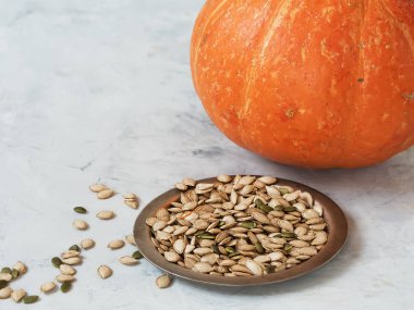 Kavanozda kabak çekirdeği var ve kasenin yanına saçılmış. Arka planda olgun bir balkabağı ve yarım balkabağı var. Yemek pişirmek. Hasat. Yakın plan. Hafif arkaplan. Cadılar Bayramı. Sezon yemeği.