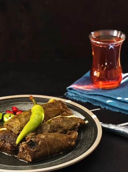 Feuilles de raisin farcies (plat méditerranéen Dolma) sur une assiette noire décorée de poivre. Sur une serviette bleue, un verre de thé. Fond en béton noir avec des stries de lumière. Vue latérale, gros plan . — Photo