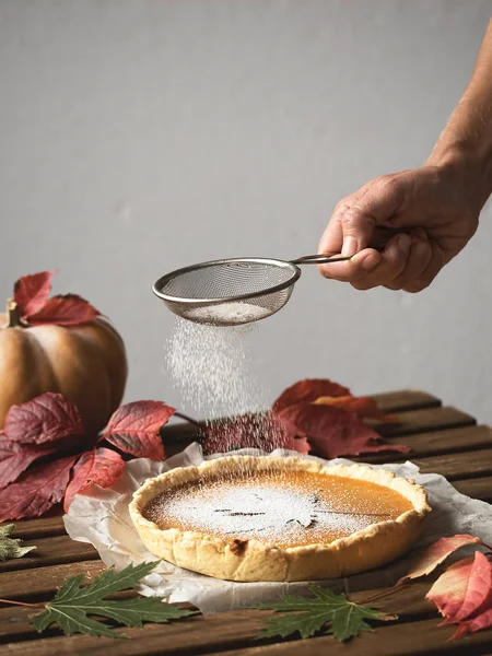 Homemade traditional American pumpkin pie located on a wooden table. In the background are colorful autumn leaves and a pumpkin. Girl decorates icing sugar cake. Top view. — Stock Photo, Image