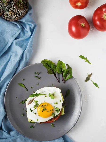 Fried egg with tomatoes and leaves of arugula and thyme on bread in a gray plate. Beautiful fried eggs. Tasty breakfast. Top view, white concrete background.