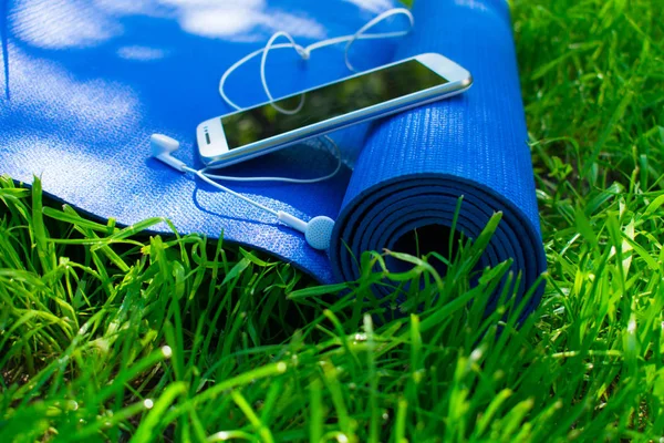A mat for yoga and pilates, a phone with headphones and on green grass, concepts of training and sports in nature