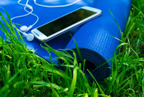 A mat for yoga and pilates, a phone with headphones and on green grass, concepts of training and sports in nature