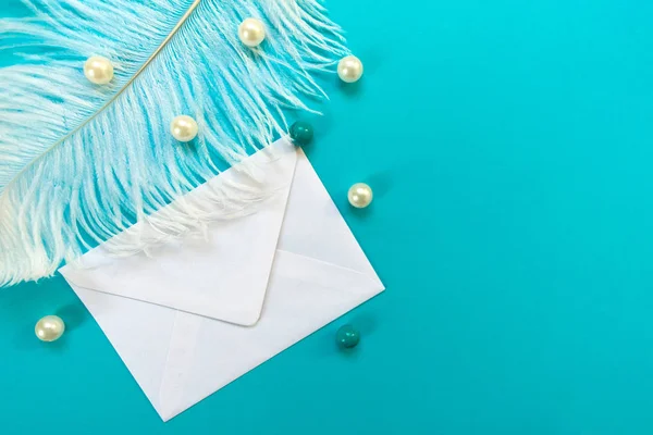 White envelope with white feather and beads isolated on blue background. Greeting card concept. Copy space.