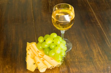 Bir kadeh beyaz şarap, dilimlenmiş peynir ve tahta bir masada bir demet üzüm.