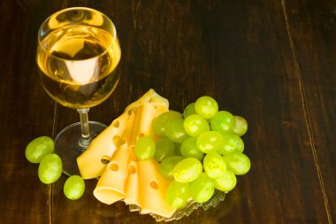 Bir kadeh beyaz şarap, dilimlenmiş peynir ve tahta bir masada bir demet üzüm.