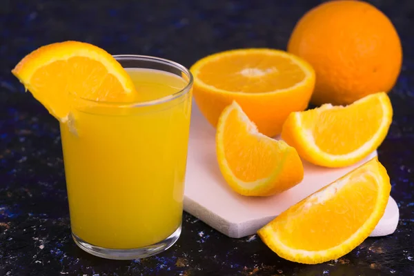 A glass of organic orange juice and sliced oranges on a dark blue background.