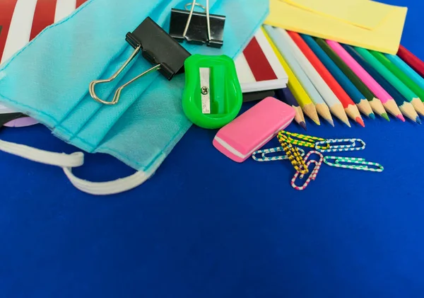 School supplies and medical mask on a blue background, returning to school after the COVID-19 pandemic. Copy space.