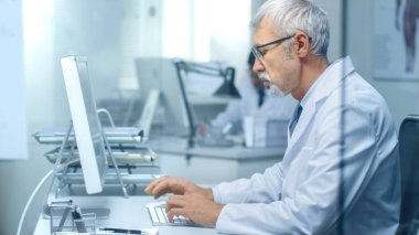 Masaüstü bilgisayarını ofisinde çalışan üst düzey tıp doktoru. Ayrıca asistanı arka planda çalışır.