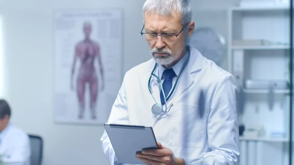 Senior Doctor Thinking About Diagnosis and Consulting Tablet Computer. His Office is Bright and Modern.