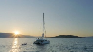 Tepenin arkasından yükselen güneş ile sakin deniz yoluyla seyahat bir yelkenli katamaran tekne hava atış.