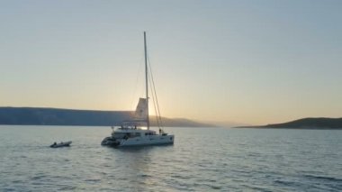 Tepenin arkasından yükselen güneş ile sakin deniz yoluyla seyahat bir yelkenli katamaran tekne hava atış.