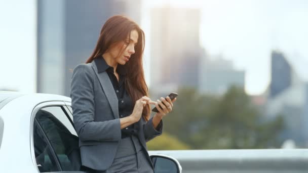 ビジネス プレミアム クラス車にもたれながらスマート フォンの用途を女性 背景の高層ビルの都会 — ストック動画