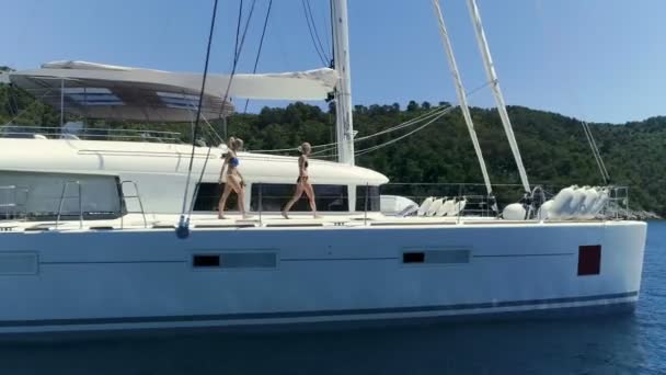 セーリング ヨットのデッキと 人の男性のスタンドアップパドル ボートの近くを歩いてビキニで 人の女性の空中アーク ショット 背景の緑の美しい海岸 — ストック動画