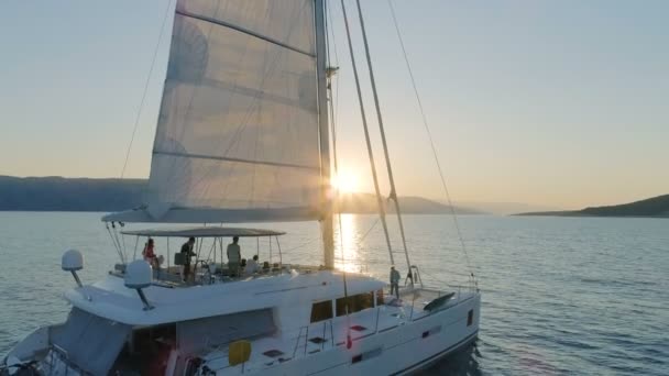 丘の背後に上昇太陽と穏やかな海を旅して上げられた帆とヨットのカタマラン ヨットの空中ショット — ストック動画