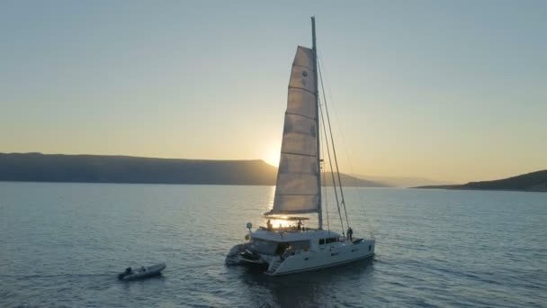 カタマラン ヨットの空中ショットは 丘の背後に上昇太陽と穏やかな海を旅して上げられた帆とヨットします 人デッキでリラックス — ストック動画