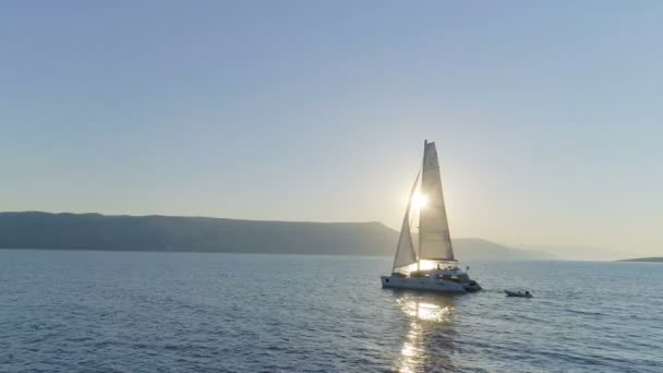 Vue Aérienne Longue D'un Bateau De Catamaran à Voile Avec Voile Levée Voyageant À Travers Les Mers Calmes Avec Soleil Se Levant Avec Collines Côtières Visibles. Les gens se relaxent sur le pont .