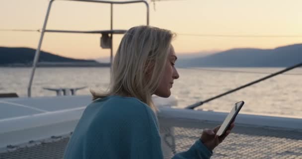 美丽的年轻女性使用智能手机 同时在游艇上放松 太阳是夕阳 岛屿在地平线上被看见 — 图库视频影像