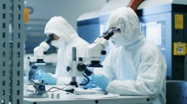 İki mühendis / bilim adamları / teknisyenleri steril temiz oda takım elbiseli kullanın mikroskoplar bileşen ayarı ve araştırma için. Onlar bir elektron komponentler fabrika üretim iş.