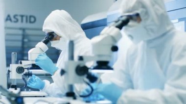 İki mühendis / bilim adamları / teknisyenleri steril temiz oda takım elbiseli kullanın mikroskoplar bileşen ayarı ve araştırma için. Onlar bir elektron komponentler fabrika üretim iş.