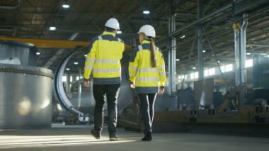 Arkadan görünüm tartışma ağır sanayi ile fabrika üretim yürürken sahip erkek ve dişi Endüstri mühendisleri bir kadeh. Büyük metal yapılar, boru hattı öğeleri etrafında yalan.
