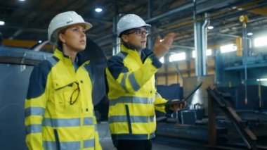 Erkek ve kadın Endüstri mühendisleri zor şapka ve güvenliği ceketler tartışmak yeni projesi kullanarak Laptop süre. Onlar ağır metal bileşenleri etrafında yalan ile fabrika üretim sanayi üzerinde yürümek-den geçerek.