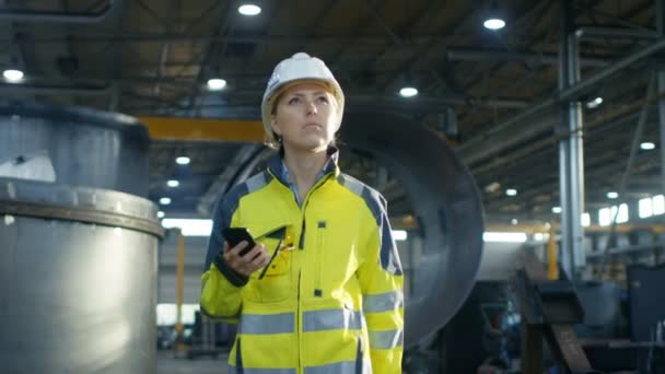 在硬帽中的女工业工人在穿过重工业制造厂时使用手机 在背景下各种金属制品工程零件的谎言 — 图库视频影像