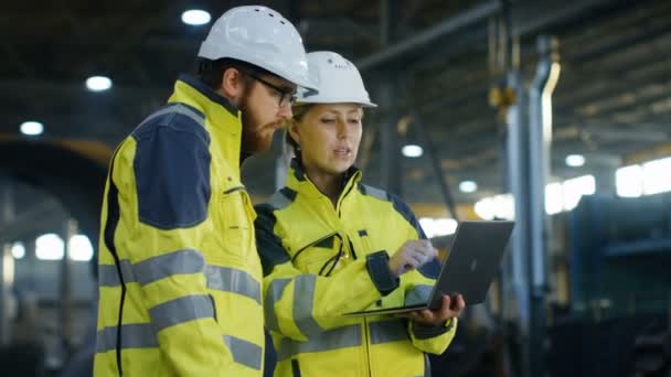Ingénieurs industriels masculins et féminins dans Hard Hats Discuter du nouveau projet tout en utilisant un ordinateur portable. Ils font des gestes. Ils travaillent à l'usine de fabrication de l'industrie lourde. Tir long .
