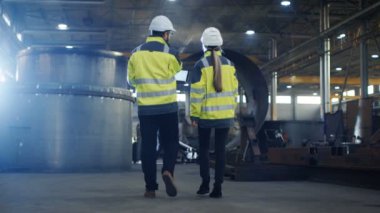 Erkek ve kadın Endüstri mühendisleri kullanımı dizüstü bilgisayar ve var tartışma iken yürüme yoluyla ağır sanayi üretim fabrikası. Sabit şapka ve emanet ceket giymek.