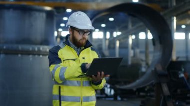 Endüstri Mühendisi sabit hat Emanet ceket kullanır dokunmatik ekran Laptop ve yürüyüşler. O ağır fabrika üretim sanayi çalışıyor. Arka plan kaynak içinde / Metal maden işleme işlemleri sürüyor.