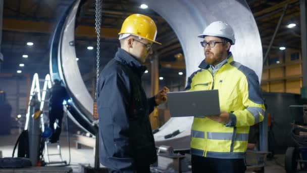À l'intérieur de l'usine industrielle lourde, l'ingénieur industriel tient un ordinateur portable et discute avec le gestionnaire de projet. Ils portent des chapeaux durs et des vestes de sécurité. En arrière-plan Soudage / Métallurgie en cours .