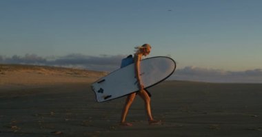 Yan görünümü görüntüleri denize doğru yürüyüş Surfboard taşıma güzel ve genç birbayan. Kız olan sarışın saç ve Backpack.The Beach boş ve güneş batıyor.