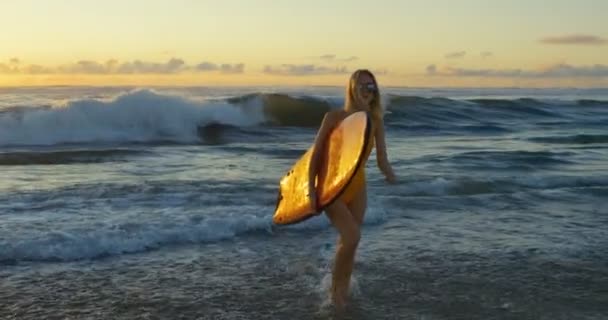 サーフボードを運ぶ中海外歩く水着で美しい若い女性 彼女はゴージャスに見える スリムがブロンドの髪です 大きな海洋波と背景の夕日 — ストック動画