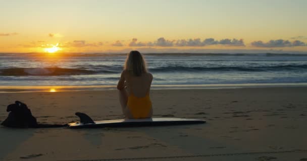 穿着泳装的美丽的年轻女子坐在海滩上 在她的冲浪板看着大洋巨浪和观看日落 圆弧拍摄素材 — 图库视频影像
