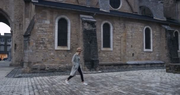 コートと帽子を身に着けているスタイリッシュな若い女性は 古いヨーロッパ教会ヤードを説明します 美しい少女と旧市街の歴史的建造物 — ストック動画
