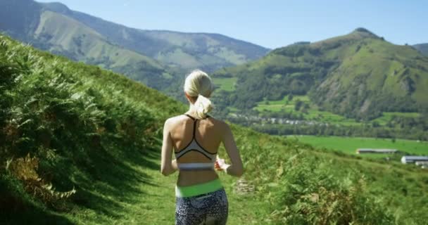 下面的影片的美丽的适合妇女赛跑运动员在运动服慢跑在小径与壮丽的山景 迷人的金发女孩和风景优美的自然 慢动作 中型射击 — 图库视频影像