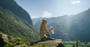 Genç Sarışın kadın spor giyim oturur dağ ve içecekler su Resting uygun. Nefes kesen tepeler, dağlar ve vadi manzaralı. Yaz saati doğal doğa manzaralı.