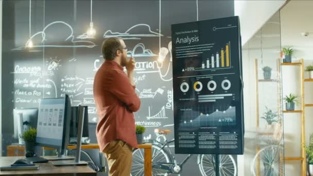 マーケティング担当者は タッチ スクリーンの統計的成長に関する最新のグラフやチャートを表示するインタラクティブなホワイト ボードに見えます 彼は創造的なオフィスで働いてください — ストック動画
