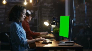 Güzel kadın ofis çalışanı vasıl onun okul sırası bir Mock-up yeşil ekran kişisel bilgisayarda çalışır. Onun iş arkadaşı oturur yanında. Yaratıcı Office akşam.
