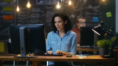 Güzel kadın ofis çalışanı masasının bir kişisel bilgisayarda çalışıyor, o onun iş ve gülümsüyor tatlı kamera için keser. Yaratıcı ofiste arka plan iş arkadaşı.