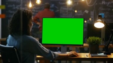 Güzel kadın ofis çalışanı vasıl onun okul sırası bir Mock-up yeşil ekran kişisel bilgisayarda çalışır. Omuz görüntüleri. Onun iş arkadaşı arka planda çalışır. Yaratıcı Office akşam.
