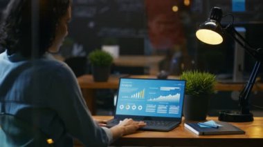 Onun masasında kadın analisti istatistikleri, grafikler ve gösterilen bir dizüstü bilgisayarda çalışır. O yaratıcı ofiste ahşap tabloda çalışıyor. Omuz görüntüleri.