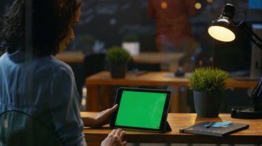 Genç kadın oturur onun ahşap masa Mock-up yeşil ile Tablet bilgisayarı kullanan ekran, o kullanır çeşitli çalmak ve dokunmatik jestleri.