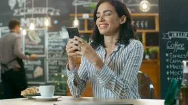 Güzel kadın Cafe kullanır Smartphone içinde oturur. Her arkadaş mesajlaşma ya da sevilen bir süre gülümsüyor. İçecek ve kruvasan ile onun tablo bardakta. Arka planda şık kahve evi meşgul.