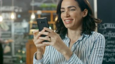 Güzel kadın Cafe kullanır Smartphone içinde oturur. Her arkadaş mesajlaşma ya da sevilen bir süre gülümsüyor. Arka plan şık kahve konut ve meşgul garson.