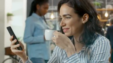 Güzel kadın Cafe kullanır Smartphone, o gülümsüyor oturur. İçecek ve kruvasan ile onun tablo bardakta. Arka planda şık kahve evi meşgul.