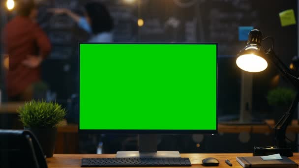 クリエイティブ オフィスの黒板壁の背景の人々 の仕事で 事務所の机の上にモックアップ緑色の画面モニターとパソコンが立っています — ストック動画