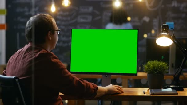 彼のデスクトップのオフィスの従業員は モックアップの緑色の画面のパソコンで動作します 肩の映像 彼の同僚は バック グラウンドで動作します クリエイティブ オフィスの夜 — ストック動画